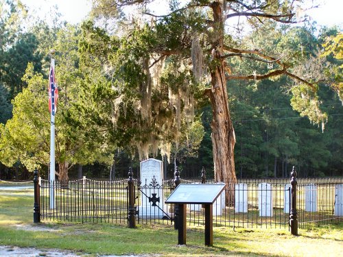 A historic cemetery with a black iron fence, gravestones, a sign, and a Confederate flag nearby, surrounded by trees.