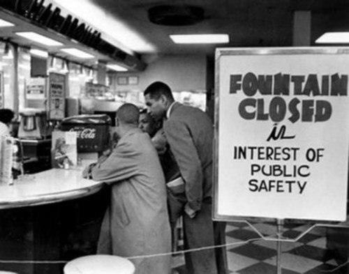A vintage diner scene with a sign reading "Fountain Closed in Interest of Public Safety" and patrons at the counter.