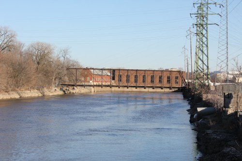 A river flows beside a brown brick building and a metal bridge, with power lines and trees lining the bank.