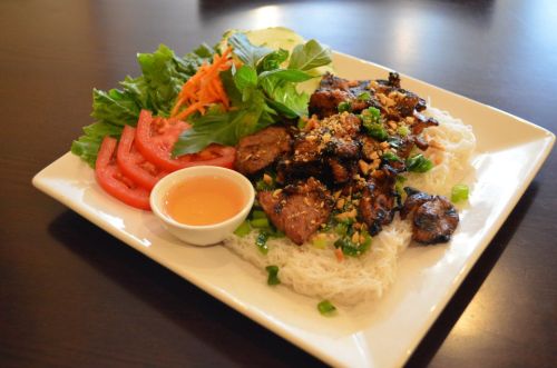 A plate of grilled meat over rice noodles, garnished with fresh herbs, vegetables, and a small bowl of dipping sauce.