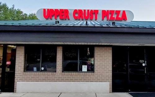 Sign for Upper Crust Pizza on a brick building, featuring large red letters and a green roof. Windows display menu items.
