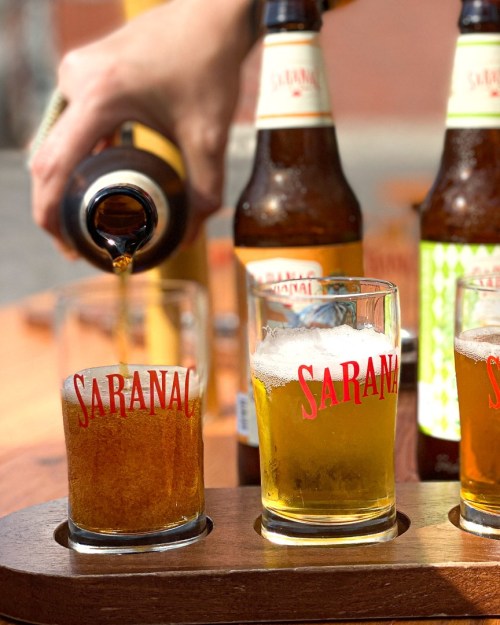 A hand pours beer into a glass labeled "SARANAC," with other bottles and glasses in the background.