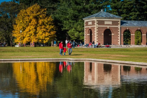 Two people in red walk by a pond, with colorful trees and a brick structure reflected in the water.