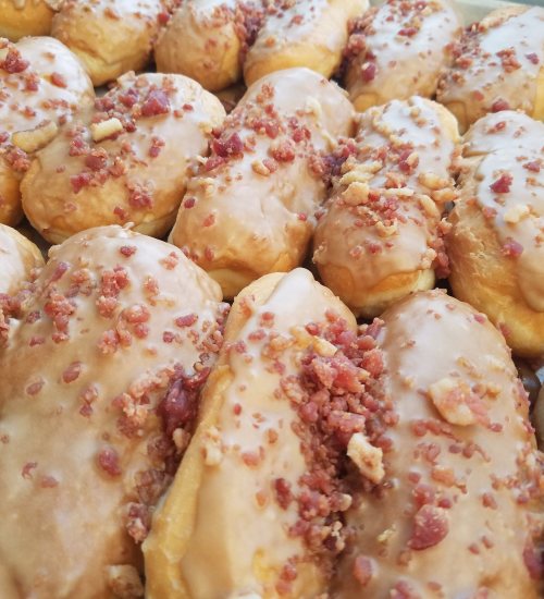 A close-up of glazed donuts topped with crispy bacon bits, arranged in a tray.