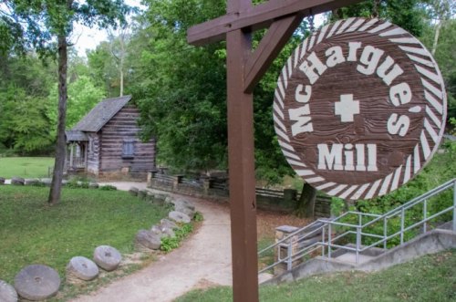 Sign for McHargue's Mill with a rustic log cabin and stone millstones in a green, wooded area.