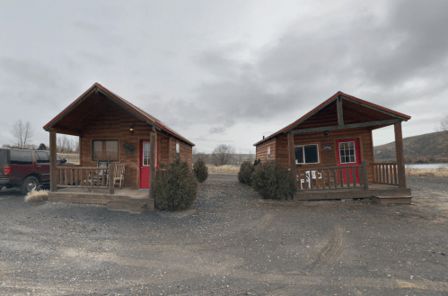 Two rustic wooden cabins with porches, set against a cloudy sky and grassy landscape.
