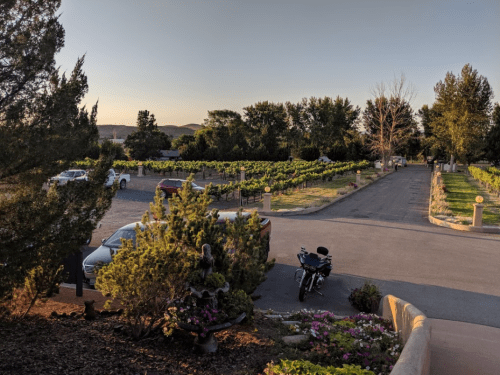 A scenic vineyard at sunset, with parked cars, trees, and a motorcycle in the foreground.