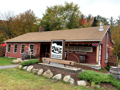 A rustic red restaurant with a sign reading "Rustic River Restaurant," surrounded by greenery and autumn foliage.