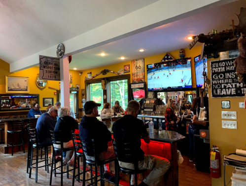 A cozy bar scene with patrons seated at the counter, watching a sports game on TV, surrounded by decor and signage.