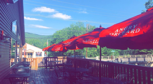 A sunny outdoor patio with red umbrellas featuring the Angry Orchard logo, surrounded by green trees and blue skies.