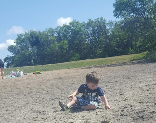 A young child sits on a sandy beach, playing, with trees and a person in the background.