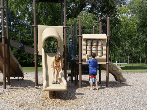 Two children play on a playground: one slides down while the other interacts with a play panel.