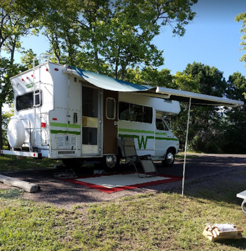A parked RV with an awning, set up on a grassy area, surrounded by trees and a small outdoor mat.