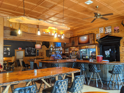 A rustic bar interior with wooden decor, a long table, and a well-stocked bar area under warm lighting.