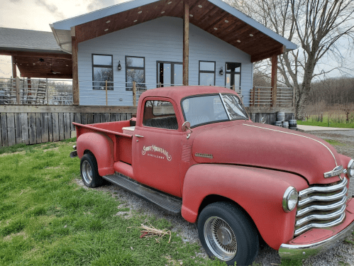 A vintage red pickup truck parked in front of a modern building with large windows and a wooden deck.