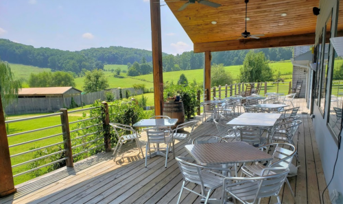 A sunny outdoor patio with metal tables and chairs, overlooking a green landscape and hills in the background.