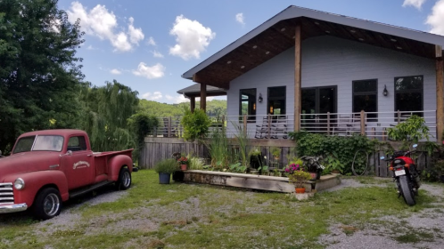 A rustic house with a porch, surrounded by greenery, featuring a vintage red truck and a motorcycle parked nearby.