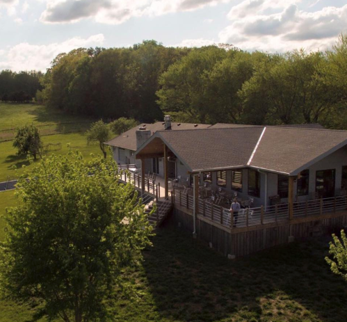 Aerial view of a modern house with a large deck, surrounded by greenery and trees under a partly cloudy sky.