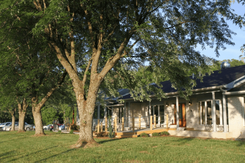 A peaceful scene of a house surrounded by trees, with a grassy area and a parking lot in the background.