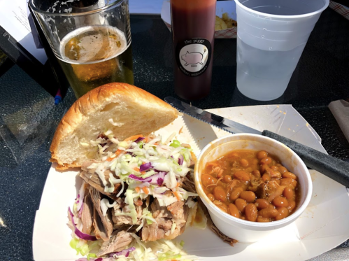 A pulled pork sandwich with coleslaw, baked beans, a beer, and a cup of water on a table.