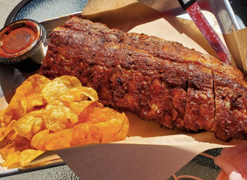A platter featuring a large, seasoned rack of ribs alongside crispy potato chips and a small container of barbecue sauce.