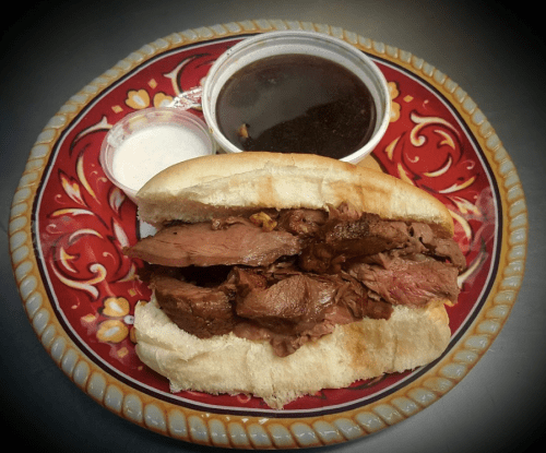 A hearty sandwich filled with sliced beef, served on a decorative plate with dipping sauce and a side of ranch dressing.