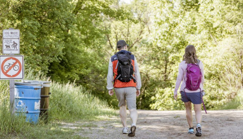 Two hikers walk along a dirt path surrounded by greenery, with signs for dog waste and no biking nearby.