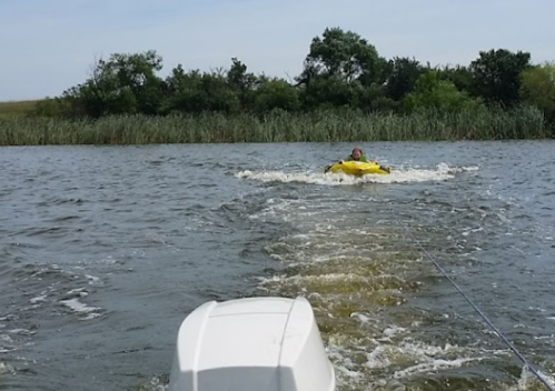 A person on a yellow inflatable tube is being towed across a calm lake, surrounded by greenery and trees.