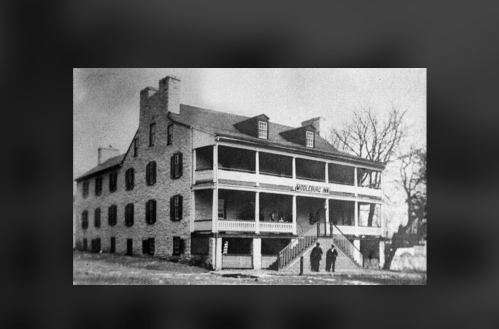 Historic black-and-white photo of a large stone building with porches, featuring people standing on the steps.