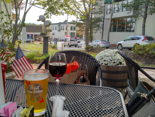 A table with a beer and wine glass, surrounded by flowers and flags, overlooking a street with cars and shops.