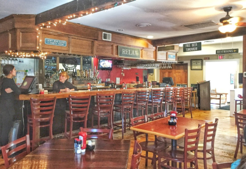 A cozy bar interior with wooden tables, chairs, and a long counter, featuring a relaxed atmosphere and warm lighting.