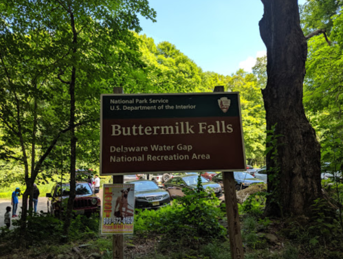 Sign for Buttermilk Falls at Delaware Water Gap National Recreation Area, surrounded by trees and parked cars.