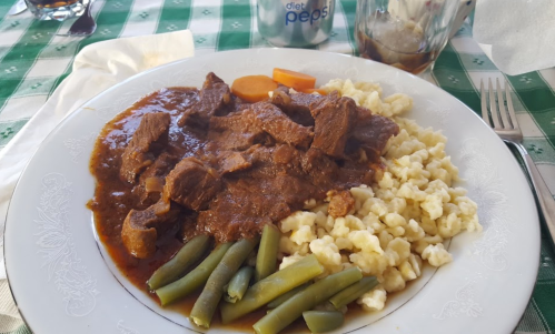 A plate of beef stew with noodles, green beans, and carrots, served on a checkered tablecloth with a drink in the background.