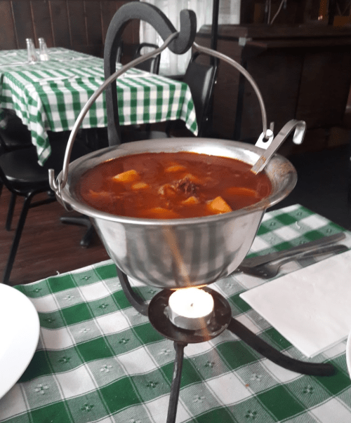 A metal pot of stew sits on a stand over a candle, with a checkered tablecloth in the background.