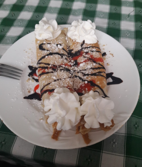 Two dessert crepes topped with whipped cream, chocolate sauce, and fruit, served on a white plate on a checkered tablecloth.