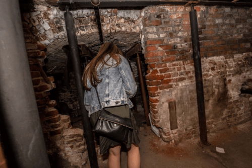 A person with long hair in a denim jacket walks through a dimly lit, brick-walled basement.