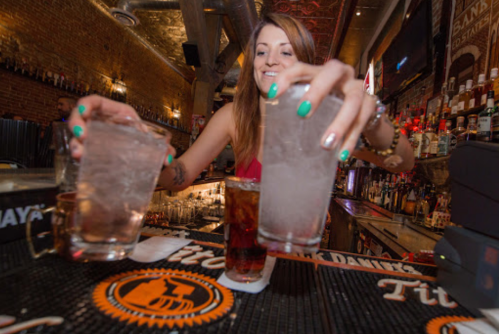 A bartender prepares drinks at a lively bar, holding two glasses filled with ice and a cocktail in front.