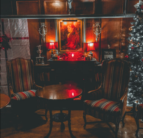 Cozy room with two striped chairs, a wooden table, a vintage portrait, and a decorated Christmas tree. Warm lighting.