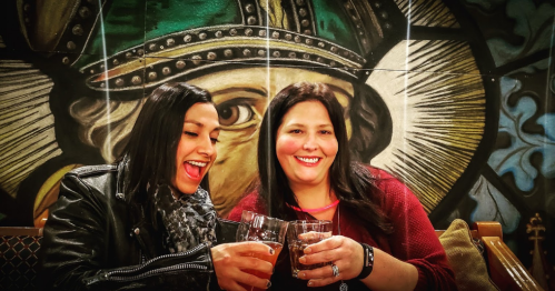 Two women clink glasses in celebration, smiling against a colorful mural backdrop.