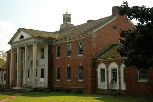 An old brick building with a columned entrance and a weathered roof, surrounded by overgrown grass and trees.