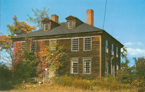 An old, two-story wooden house with ivy, surrounded by overgrown grass and trees under a clear blue sky.