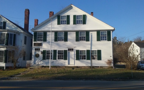 A white, two-story house with green shutters and a front porch, set against a clear blue sky.