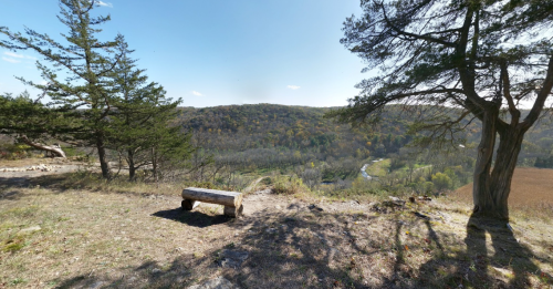 Scenic view of a valley with trees and a log bench overlooking a winding river and rolling hills under a clear sky.