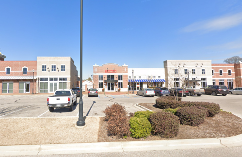 A shopping plaza with several storefronts, parked cars, and a clear blue sky in the background.