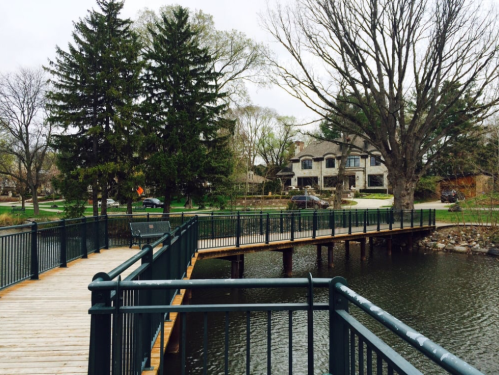 A wooden walkway extends over a calm lake, surrounded by trees and a residential area in the background.