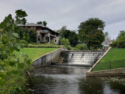 A serene scene featuring a small waterfall flowing into a calm river, surrounded by green grass and trees, with buildings nearby.