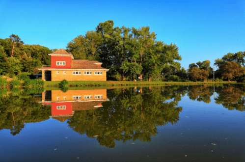A serene lake scene with a house reflecting in the water, surrounded by lush greenery and a clear blue sky.