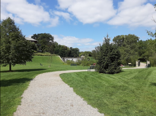 A winding gravel path through a green park with trees and a distant stone wall under a partly cloudy sky.