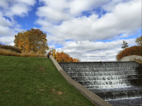 A person stands on a grassy slope beside a waterfall, with colorful autumn trees and a cloudy sky in the background.
