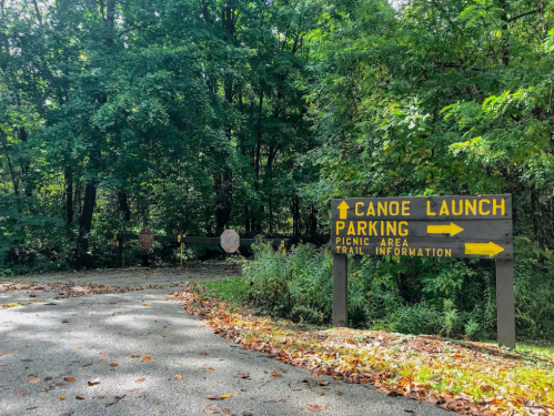 Signpost at a wooded area indicating directions to canoe launch, parking, picnic area, and trail information.